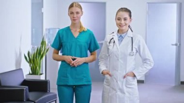 Kameranın önünde güzel doktor ve hemşire asistanı sarışın bayan kameranın önünde duruyor ve hastane koridorunda gülümsüyor.
