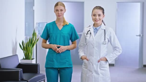 Przed kamerą piękna pani doktor i jej asystentka pielęgniarki blondynka stojąca przed kamerą i uśmiechnięta w szpitalnym korytarzu. — Wideo stockowe