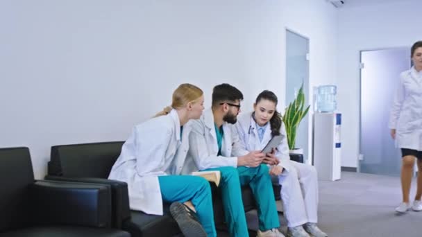 Groep medische studenten hebben praktijkles ze zitten op de bank in de ziekenhuisgang en analyseren de diagnose van de patiënt met behulp van digitale tablet andere artsen lopen rond — Stockvideo
