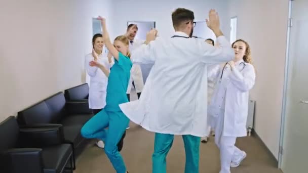 В современном коридоре в больнице группа медицинского персонала врачи и медсестры танцуют и чувствуют возбуждение в конце рабочего дня — стоковое видео