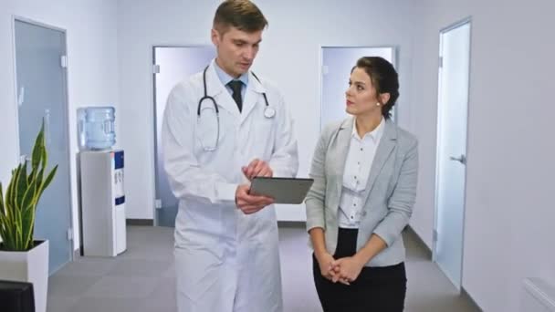 Nowoczesny korytarz szpitalny piękna dojrzała pacjentka i dojrzały lekarz rozmawiający o przyszłym leczeniu podczas spaceru korytarzem — Wideo stockowe
