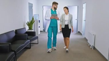 Hastanenin koridorunda elinde dijital tabletle yakışıklı bir doktor ve koridordan yürürken kan testinin sonuçlarını analiz eden olgun bir kadın var.