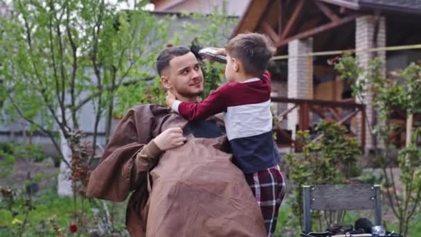 Zabawny czas mały ładny chłopiec próbuje zrobić fryzurę dla swojego starszego brata w ogrodzie, podczas gdy siedzi w ogrodzie. 4k — Wideo stockowe