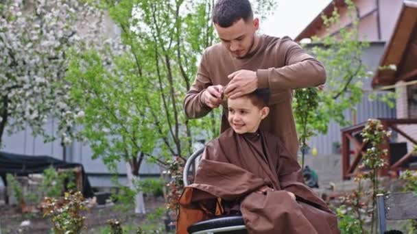 Taglio di capelli tempo in giardino carismatico bambino rimanere sulla sedia e fare un sorriso mentre l'uomo barbiere fare il taglio di capelli — Video Stock