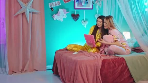 Toll aussehende Damen plaudern zusammen im Schlafanzug auf dem Schlafzimmer, sie haben eine Mädchennacht mit einem Tablet, das etwas beobachtet — Stockvideo
