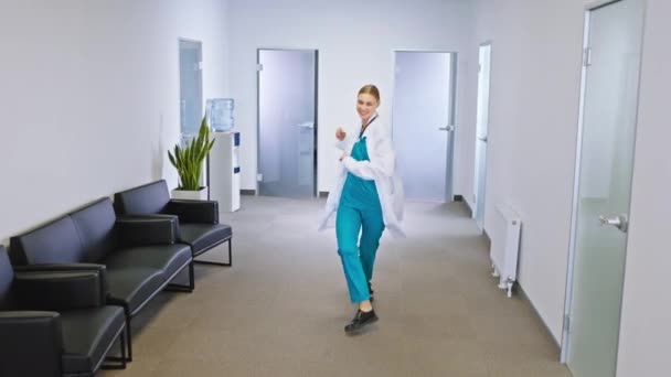 Feliz y joven doctora de familia bailando delante de la cámara en el pasillo del hospital ella está tan emocionada. v) — Vídeos de Stock