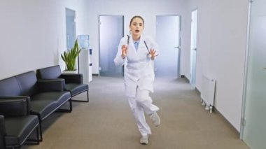 Gülümseyen genç doktor hanım kameranın önünde heyecanla dans ediyor ve iş yerinde eğleniyor. ARRI Alexa Mini 'de çekilen