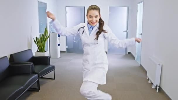 Modern hastane koridorunun ortasında kameranın önünde dans eden çok heyecanlı bir kadın doktor. — Stok video