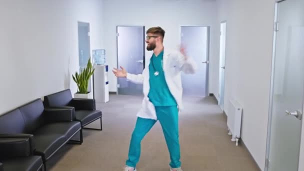 Voor de camera charismatische man verpleegster met een grote glimlach dansen gelukkig in het midden van de gang van het ziekenhuis hij zijn erg enthousiast — Stockvideo
