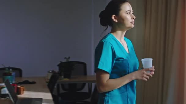 在一家现代化的医院里，一位迷人的护士女士来到全景窗口，喝了些咖啡，在客房里享受她的休息时间 — 图库视频影像