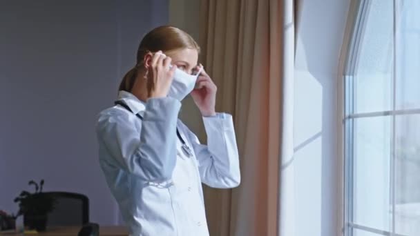 Piękna młoda pani doktor wziąć jej maskę ochronną i noszenie na twarzy i patrząc smutne w panoramiczne okno — Wideo stockowe