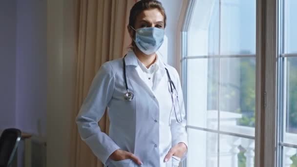 Belle femme médecin avec un masque de protection à l'hôpital regardant par la fenêtre panoramique et ont un visage fatigué dans sa chambre de bureau — Video