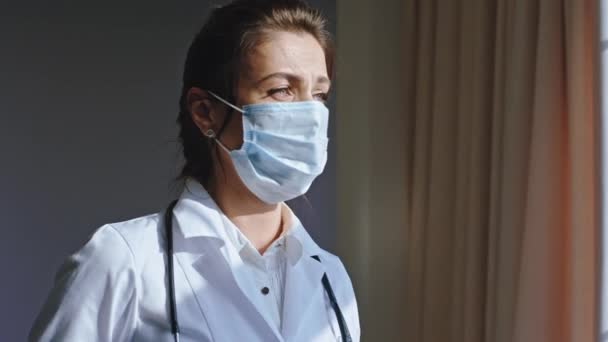 Modern hospital in the stuff room portrait of a tired and sad woman doctor wearing a protective mask she looking through the window Coronavirus pandemic 2019 — Stock Video