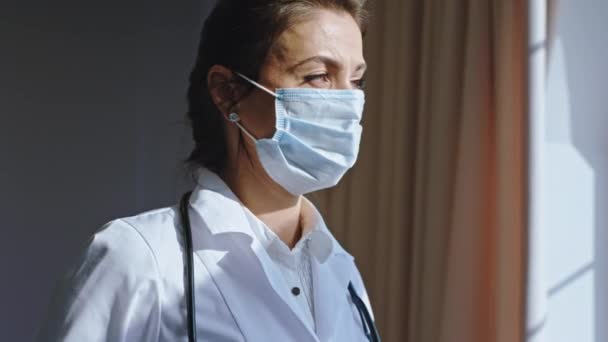Ώριμη όμορφη γυναίκα με προστατευτική μάσκα κάνει ένα διάλειμμα στο δωμάτιο του γραφείου της κοιτάζει μέσα από το παράθυρο του νοσοκομείου με πανοραμική θέα — Αρχείο Βίντεο