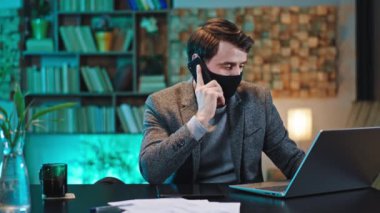 Modern ofis çalışanı koruyucu maske takıyor telefonda konuşuyor ve Coronavirus 2019 dizüstü bilgisayarına bazı notlar alıyor.