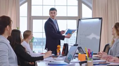 İyi görünümlü beyaz iş adamı iş arkadaşlarına yeni bir proje planı sunuyor. Fikirleri açıklıyor ve beyaz tahtada bazı istatistikler gösteriyor.
