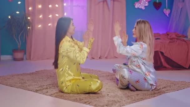 Bästa vänner damer i en vacker pyjamas har en bra tid tillsammans på en övernattning i sovrummet de leker med händerna ner på golvet njuter av tiden — Stockvideo