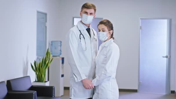 Nel corridoio dell'ospedale coppia medici uomini e donne con una maschera protettiva sconvolto abbracciarsi guardano dritto alla telecamera — Video Stock