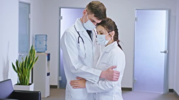 In the m of the luminous hospital corridor couple of doctors with a protective mask mask in front of the camera hugging each other they have a sad face — Stock Video