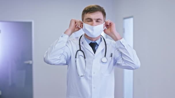 Врач носит защитную маску в современном коридоре больницы и смотрит прямо в камеру у него серьезное лицо — стоковое видео