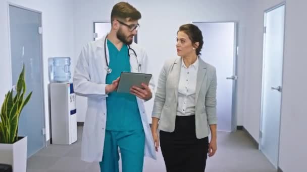 Gut aussehende Patientinnen im Krankenhaus unterhalten sich nach der Gesundheitsprüfung auf dem Krankenhausflur mit dem Arzt und werfen einen Blick in das digitale Tablet. Schuss auf ARRI — Stockvideo