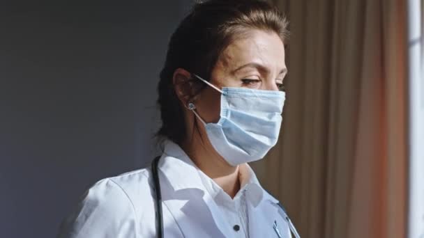 Carismática doctora con los ojos cansados y una máscara protectora mirando por la ventana del hospital cerca de la cámara — Vídeo de stock