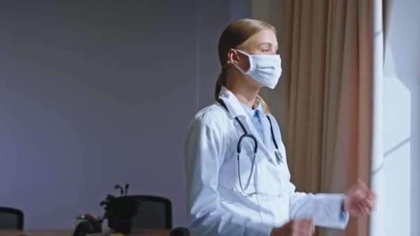 Ελκυστική νοσοκόμα κυρία με προστατευτική μάσκα κάνει ένα διάλειμμα έχει ένα σκεπτόμενο πρόσωπο κοιτάζοντας μέσα από το πανοραμικό παράθυρο και να πάρει την προστατευτική μάσκα από Covid-19 — Αρχείο Βίντεο