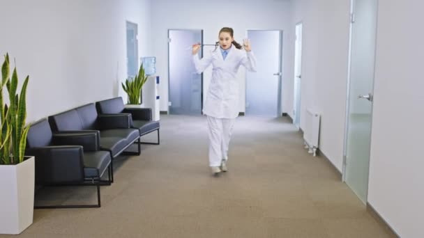 Carismática doctora con una gran sonrisa bailando emocionada en el moderno pasillo del hospital que mira directamente a la cámara. 4k — Vídeos de Stock