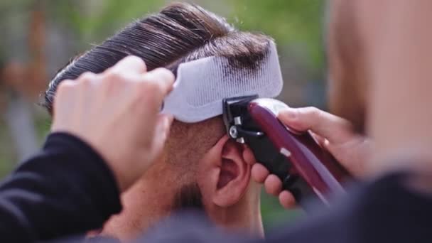 Details close-up in de tuin thuis kapper man doet een kapsel gedaan om een jonge man met een zwarte kin — Stockvideo