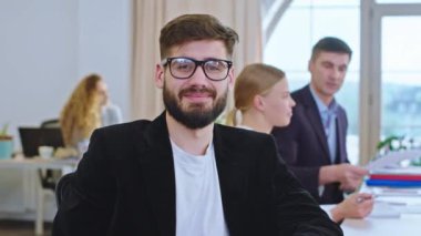 Toplantı odasında kameranın önünde genç adam doğrudan kameraya bakıyor ve gülümsüyor.