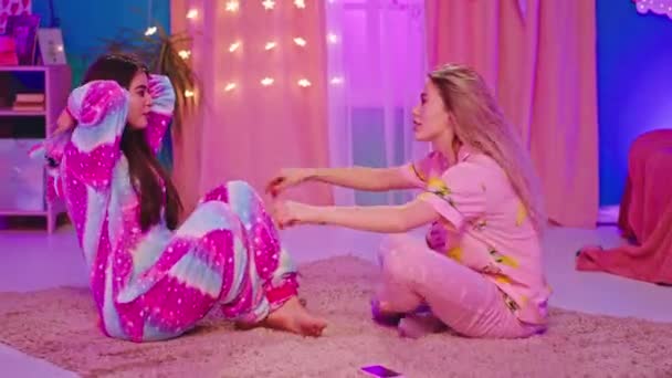 Amigos divertidos dos dos hermosas damas en un pijama divertido uno con unicornio disfrutando del tiempo juntos que se sientan en la alfombra — Vídeo de stock