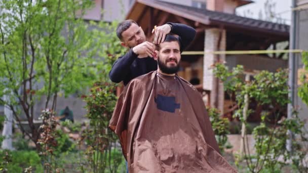 W ogrodzie w domu charyzmatyczny mężczyzna ma fryzurę, siedzi na krześle, a fryzjer robi fryzurę. — Wideo stockowe