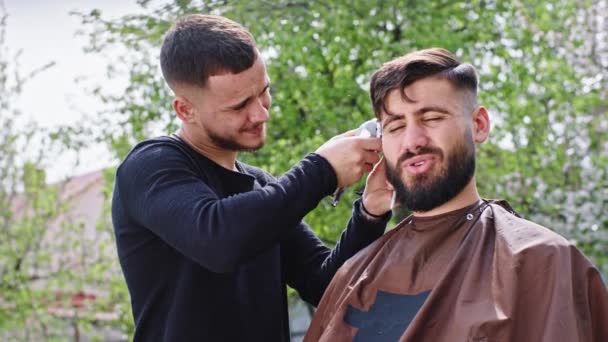 Devant la caméra, un coiffeur fait une coupe de cheveux professionnelle à son ami à la maison dans le jardin, ils sourient grand et discutent — Video