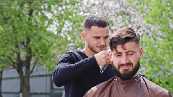Nel grande giardino verde barbiere uomo facendo un taglio di capelli al suo amico di fronte alla telecamera discutono insieme durante l'utilizzo del tagliacapelli — Video Stock