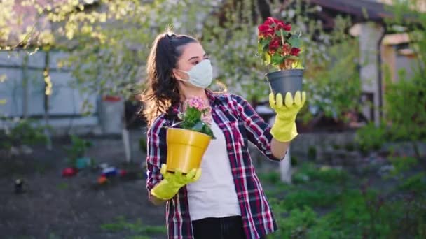 Uprostřed zahrady doma dobře vypadající mladá žena se dvěma květináči a ochrannou maskou hledící přímo do kamery a usmívající se velkou koncepcí Coronavirus 2019 — Stock video