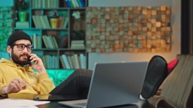Kameranın önündeki yakışıklı genç adam evde bilgisayarla çalışırken rahatlamış telefonuyla konuşuyor ve evde eğleniyor. ARRI Alexa Mini 'de çekilen