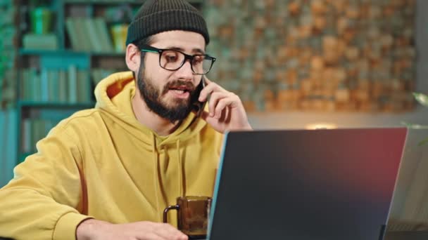 Харизматичный молодой человек разговаривает по телефону, сидя перед ноутбуком в своем домашнем офисе он работает из дома — стоковое видео