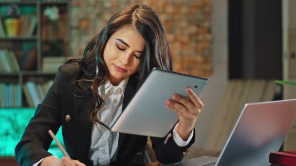 Красивая женщина в офисе работает сконцентрирована с ноутбука и планшета в то же время она печатает некоторые документы — стоковое видео