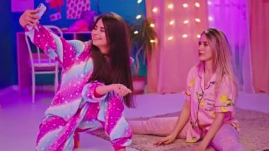Güzel bir yatak odasında iki karizmatik kadın güzel pijama giyer biri tek boynuzlu at giyer ve akıllı telefonla selfie çekerler.