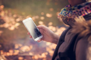 Park, lake ve yansımaları yumuşak odak rüya gibi göz ile arka planda yürürken bir smartphone tutan güzel bir esmer kadın close-up. Teknoloji ve modern yaşam kavramı.