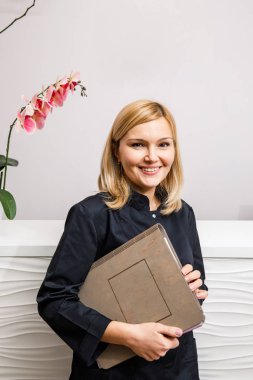 Bir güzellik salonunun siyah elbiseli resepsiyonisti beyaz bir resepsiyonun yanında duruyor.