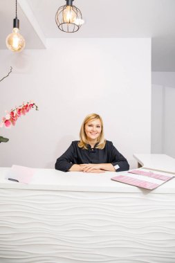 Bir güzellik salonunun siyah elbiseli resepsiyonisti beyaz bir resepsiyonun yanında duruyor.