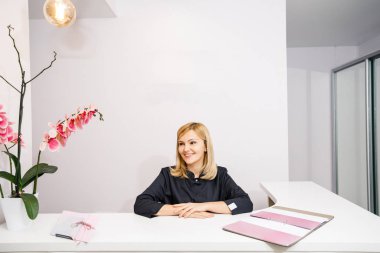 Bir güzellik salonunun siyah elbiseli resepsiyonisti beyaz bir resepsiyonun yanında duruyor.