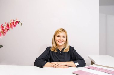 Bir güzellik salonunun siyah elbiseli resepsiyonisti beyaz bir resepsiyonun yanında duruyor.