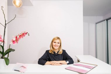 Bir güzellik salonunun siyah elbiseli resepsiyonisti beyaz bir resepsiyonun yanında duruyor.