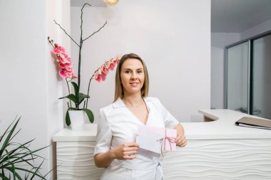 Beyaz bornozlu çekici genç bir doktor elinde hediye kartları ve güzellik salonunun arka planında duruyor.