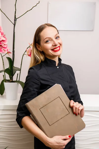 smiling young doctor in black bathrobe holding book and standing on beauty salon background