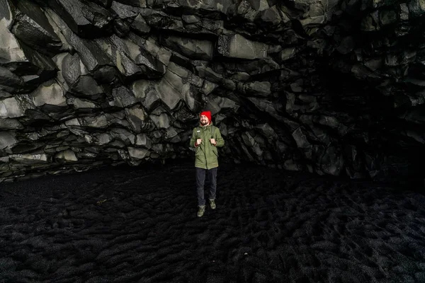 Turista Zelené Bundě Červený Klobouk Chůze Černém Písku Horské Jeskyni — Stock fotografie