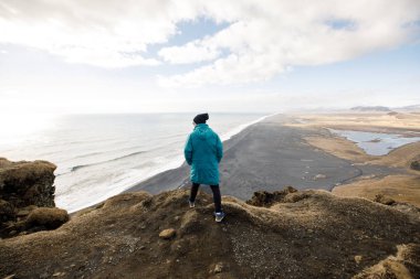 cute young man sitting on cliff with black sand beach on mountains view background