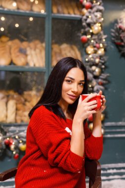 Genç, güzel, esmer, kırmızı kazaklı ve beyaz pantolonlu bir kız Paris 'te bir sokakta bir ekmek dükkanının arka planında çay içiyor.